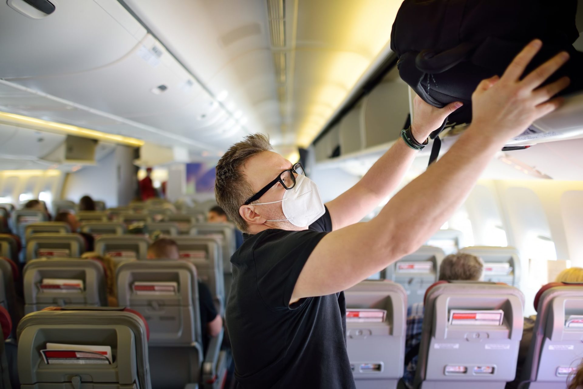 Un pasajero con mascarilla, colocando su maleta en el compartimento de un avión.