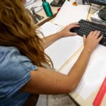 Una mujer escribe en el teclado de su ordenador, con papeles alrededor mientras trabaja en la oficina.